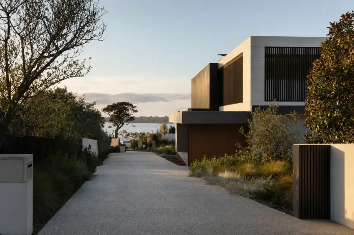 Wide honed concrete driveway leading to a modern Applecross river-frontage home, Swan River in distance, manicured landscape-architect garden with native grasses, late afternoon golden light