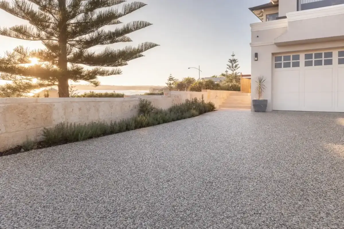 Newly poured exposed aggregate concrete driveway at a Cottesloe coastal home, sealed pebble texture in cream and grey tones, salt air haze, late golden afternoon light