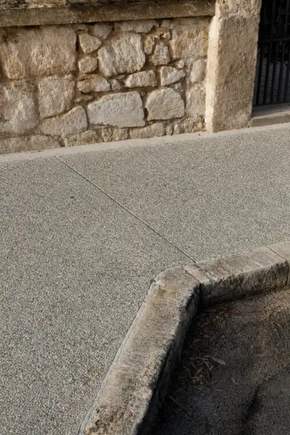 Close detail of a heritage-grey exposed aggregate concrete crossover meeting an old kerb in central Fremantle, weathered limestone wall behind
