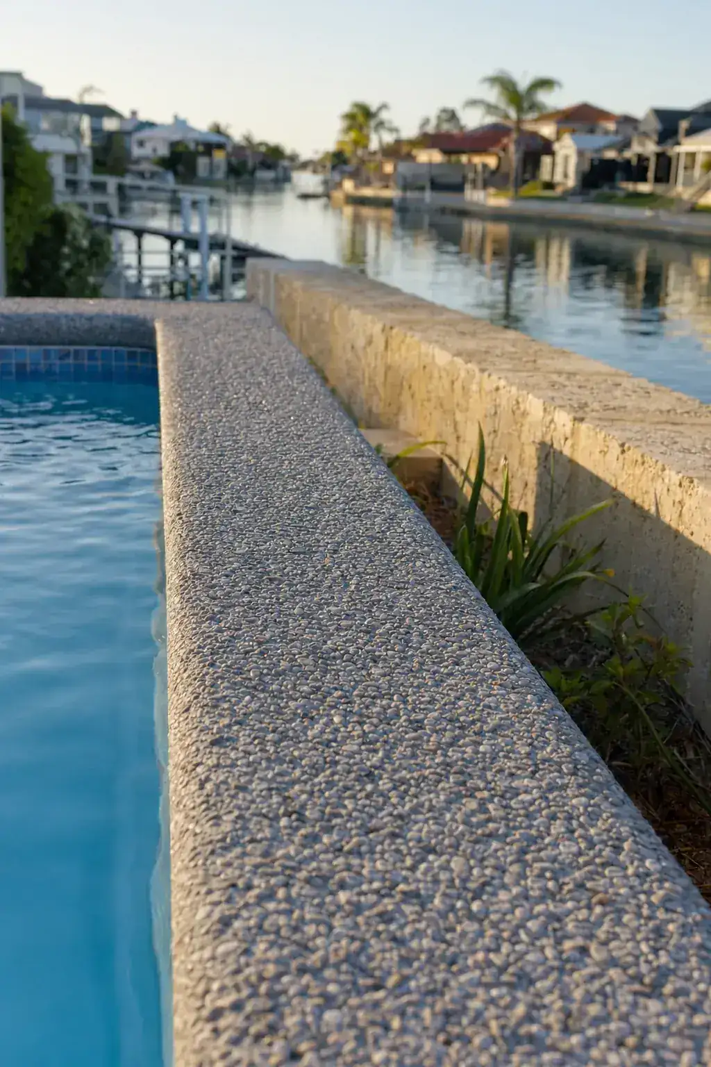 Close detail of an exposed aggregate concrete pool surround edge beside a Mandurah canal-estate property with low limestone retaining wall and calm water reflection