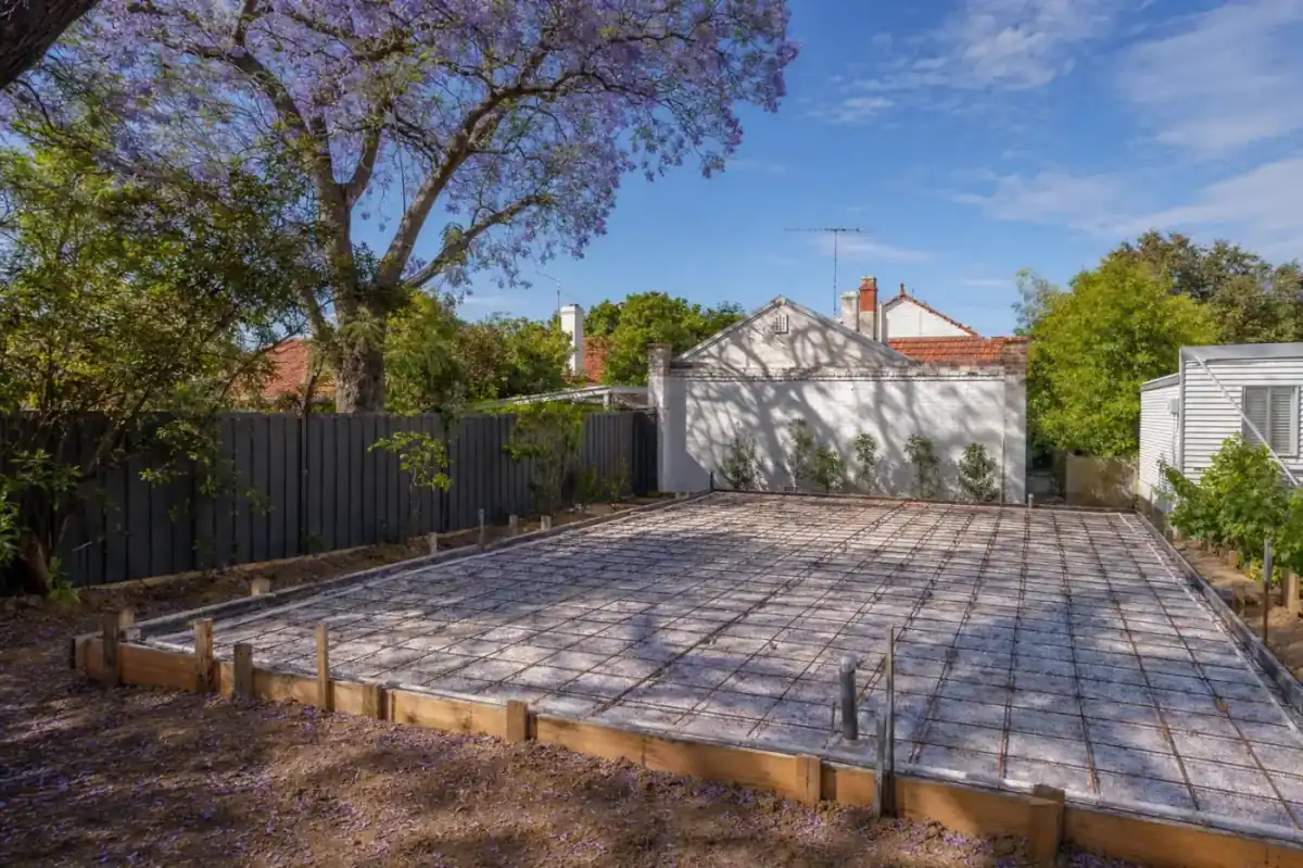 Newly poured concrete granny-flat slab at the rear of a Mount Lawley character cottage block, timber formwork still in place, mature jacaranda casting purple bloom shadow