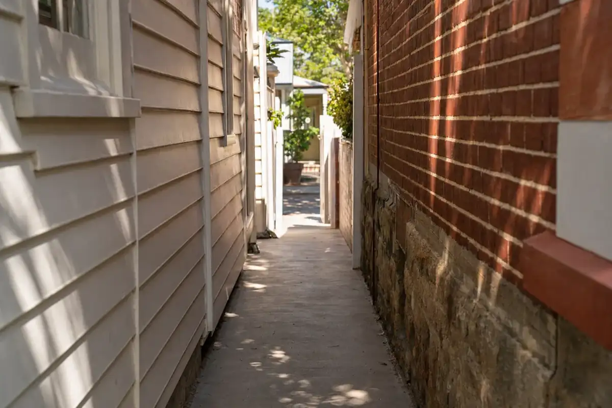 Newly poured concrete side passage path between two heritage character cottages in Subiaco, painted weatherboard wall on left, red-brick wall on right, dappled late afternoon shadow